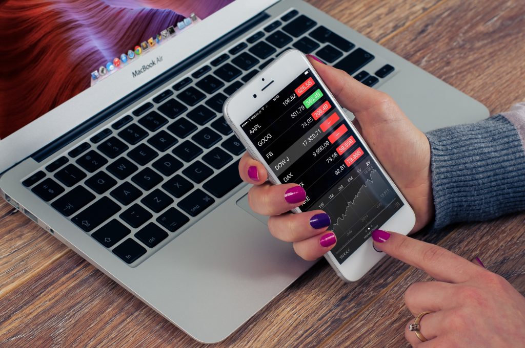 Mobile phone being held by a hand in front of a MacBook air, with pink and purple nail varnish, showing stock market figures. 